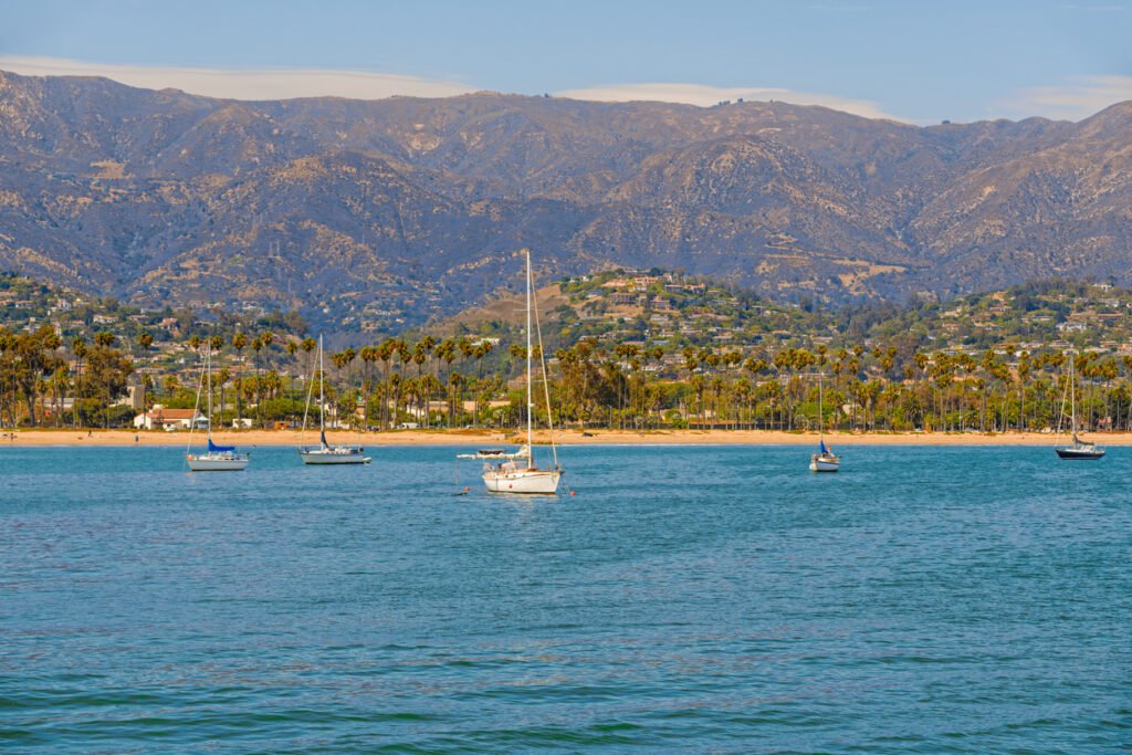 Sailboats gracefully dot Santa Barbara Harbor's shimmering waters, framed by a picturesque beach lined with swaying palm trees and embraced by majestic mountains on the horizon.